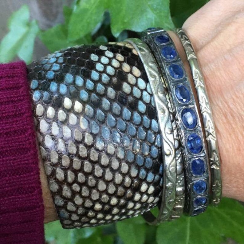 May include: A stack of bracelets on a wrist. The bottom bracelet is made of brown leather with a blue and white pattern. The other two bracelets are silver with blue gemstones.