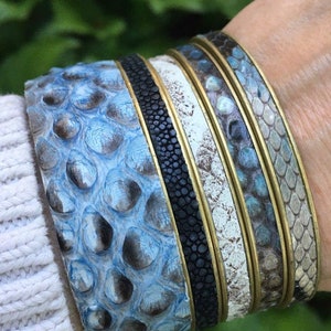 K&ouml;nnte beinhalten: Stapel von vier Armb&auml;ndern aus blauem, wei&szlig;em und schwarzem Leder mit Messingakzenten. Die Armb&auml;nder haben ein strukturiertes, schuppiges Muster.