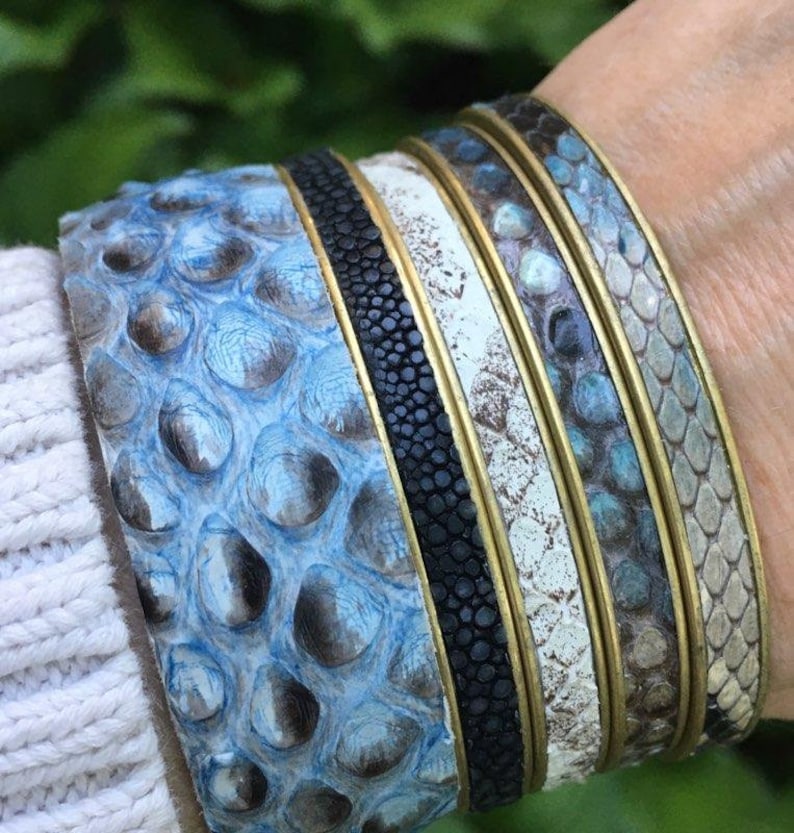 K&ouml;nnte beinhalten: Stapel von vier Armb&auml;ndern aus blauem, wei&szlig;em und schwarzem Leder mit Messingakzenten. Die Armb&auml;nder haben ein strukturiertes, schuppiges Muster.