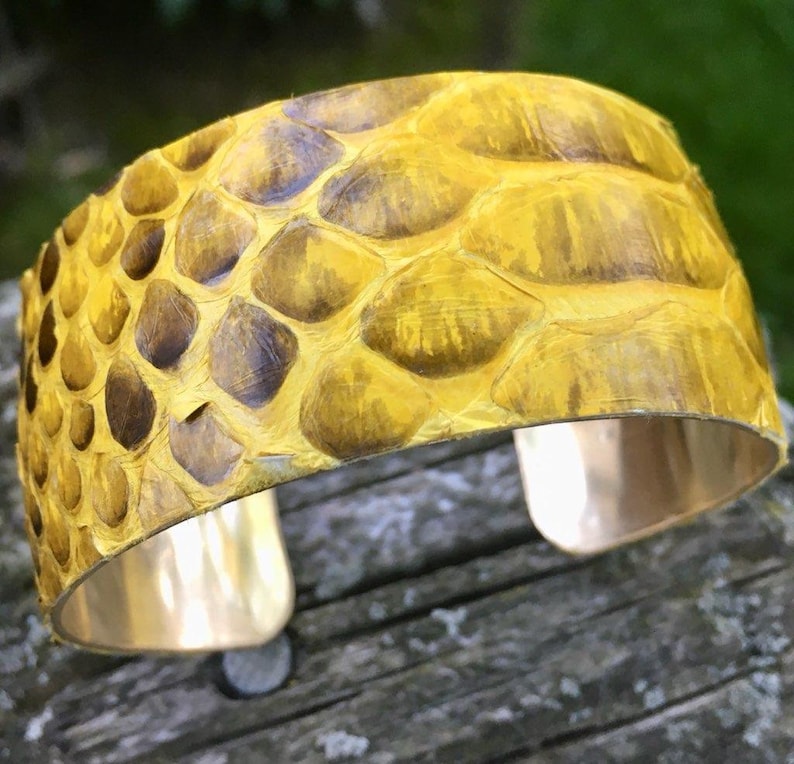 May include: A yellow snakeskin cuff bracelet with a gold metal base. The bracelet is made of real snakeskin and has a unique, textured look.