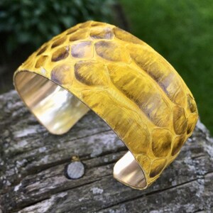 Könnte beinhalten: Ein gelbes Lederarmband mit Schlangenhautmuster. Das Armband ist aus Messing und hat eine polierte Oberfläche.