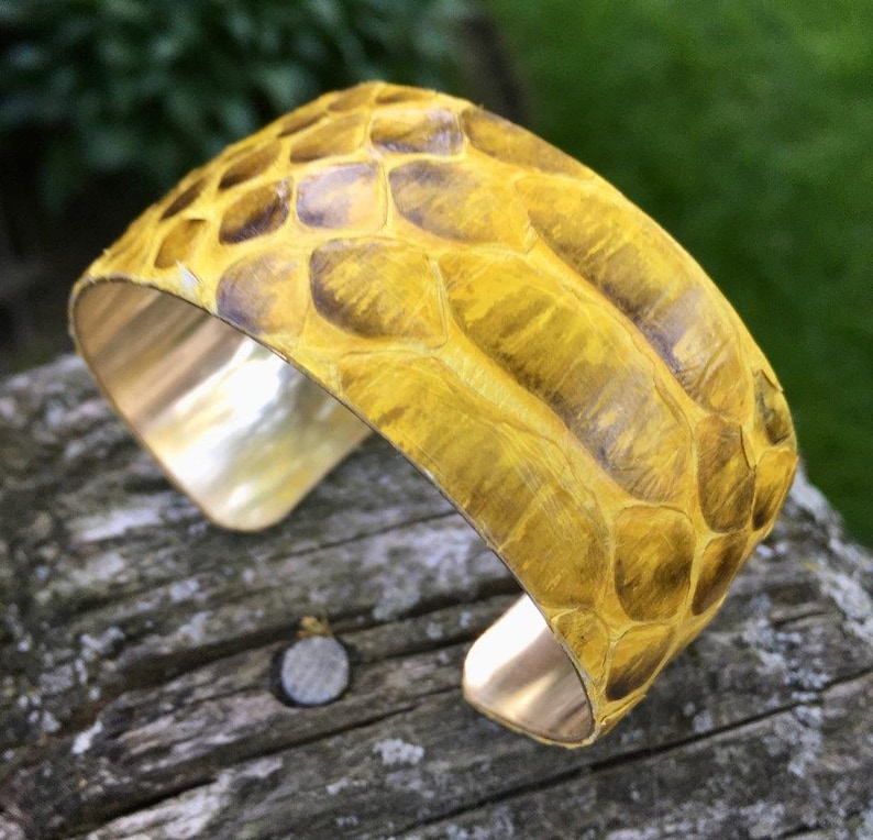 May include: A yellow leather cuff bracelet with a snakeskin pattern. The bracelet is made of brass and has a polished finish.