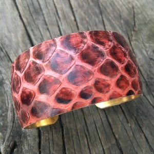 Könnte beinhalten: Ein rotes Lederarmband mit Schlangenhautmuster. Das Armband ist aus echtem Leder und hat einen Messingverschluss.