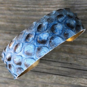 K&ouml;nnte beinhalten: Ein blaues und graues Lederarmband mit einem strukturierten Muster. Das Armband ist aus echtem Leder und hat eine Messingbasis.