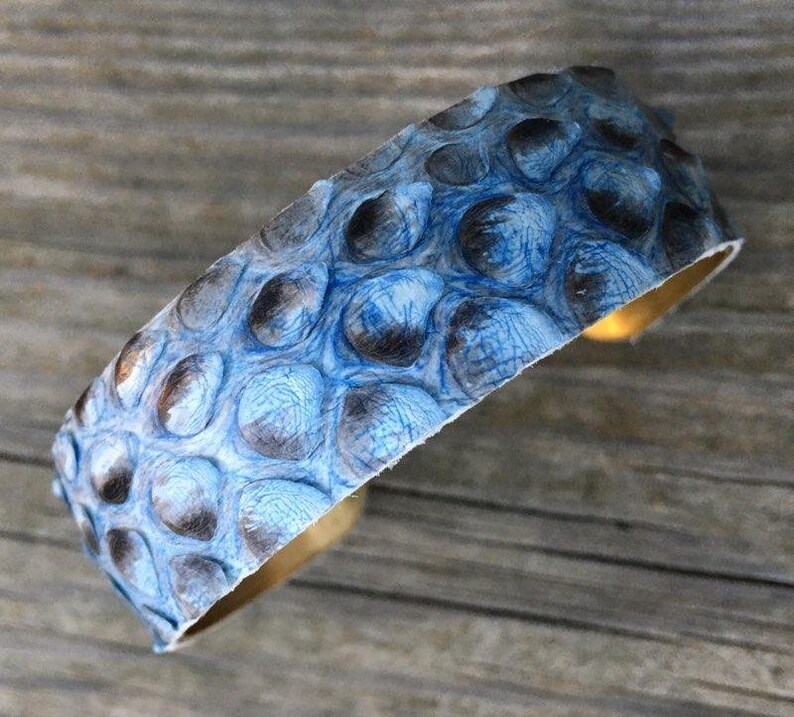 K&ouml;nnte beinhalten: Ein blaues und graues Lederarmband mit einem strukturierten Muster. Das Armband ist aus echtem Leder und hat eine Messingbasis.