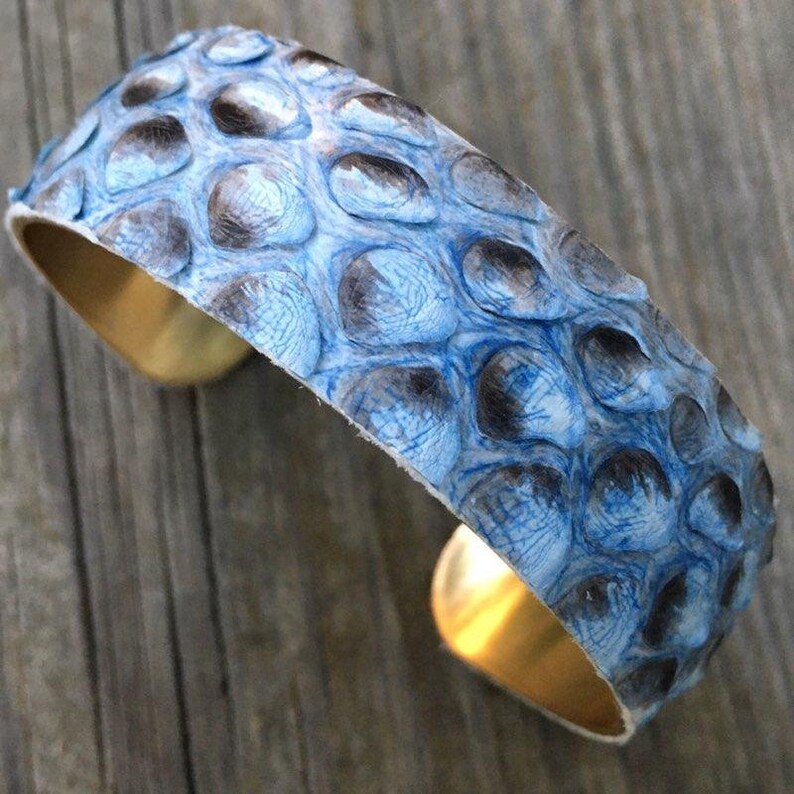 K&ouml;nnte beinhalten: Ein blaues und schwarzes Schlangenleder-Armband mit einem goldenen Metallband.