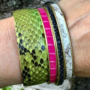 Pu&ograve; includere: Un set di quattro braccialetti impilati su un polso. I braccialetti sono realizzati con materiali diversi, tra cui pelle, metallo e tessuto. Il braccialetto superiore &egrave; in pelle verde con un motivo a pelle di serpente. Il secondo braccialetto &egrave; in tessuto rosa acceso con una texture liscia. Il terzo braccialetto &egrave; in pelle nera con una superficie strutturata. Il braccialetto inferiore &egrave; in pelle bianca con un aspetto invecchiato.