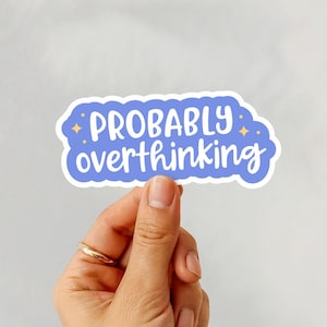 Puede incluir: Una pegatina con el texto "PROBABLY overthinking" en escritura blanca sobre un fondo azul con un borde blanco. La pegatina est&aacute; sujeta por una mano con un anillo dorado.