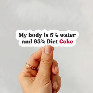 May include: A white sticker with the text "My body is 5% water and 95% Diet Coke" in black and red font. The sticker is held by a hand with a gold ring.