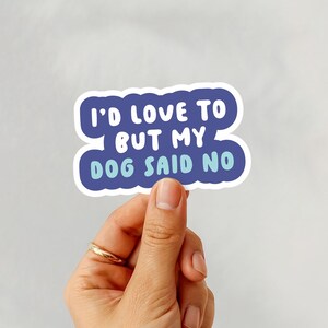 May include: A white sticker with blue text that reads "I'D LOVE TO BUT MY DOG SAID NO." The text is in a playful font and is outlined in white. The sticker is held by a hand with a gold ring.