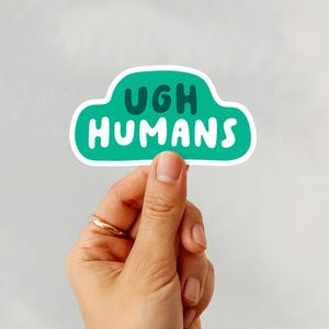May include: A sticker with the words "UGH HUMANS" in white, inside a green cloud-shaped border. The sticker is held by a hand with a gold ring, against a white background.