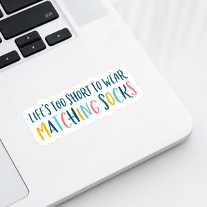 May include: A white sticker with the text "LIFE'S TOO SHORT TO WEAR MATCHING SOCKS" in colorful, playful lettering. The sticker is placed on a white laptop, with the keyboard visible in the background.