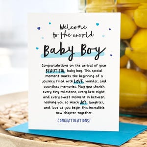 May include: A white greeting card with blue accents and text that reads "Welcome to the world Baby Boy." The card is on a woven tray with a blue envelope. The card's message congratulates the arrival of a baby boy.