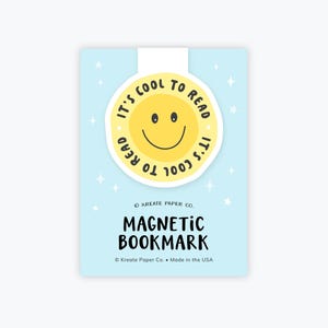 May include: A light blue magnetic bookmark with a yellow smiley face that says "It's cool to read". The bookmark also has the words "Magnetic Bookmark" in black text. The brand name "Kreate Paper Co." is also visible.