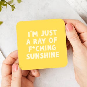 May include: A square, yellow coaster with rounded corners is held by two hands. The coaster has the text "I'M JUST A RAY OF F*CKING SUNSHINE" in white, bold letters. The background includes a ruler, a pen, and greenery.
