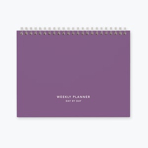 May include: Purple weekly planner with a silver coil binding and the text "WEEKLY PLANNER DAY BY DAY" printed on the front cover.