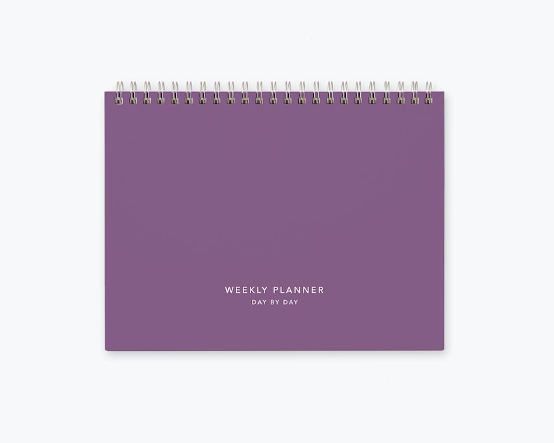 May include: Purple weekly planner with a silver coil binding and the text "WEEKLY PLANNER DAY BY DAY" printed on the front cover.