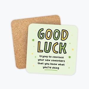 May include: A square coaster with the words "GOOD LUCK" in green and yellow, with a humorous message below. A second cork coaster is visible in the background. The coaster is a fun gift for a new coworker.