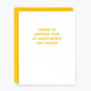 May include: A white greeting card with a yellow envelope. The card features the text "Cheers to another year of questionable life choices" in yellow. The card is a humorous gift for a friend or family member.