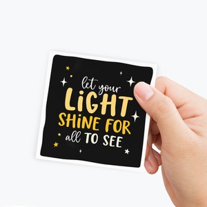 May include: A black square sticker with white and yellow text that reads "Let your light shine for all to see". The sticker has small white stars around the text.