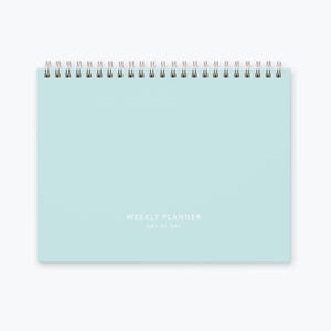May include: A light blue weekly planner with a silver coil binding. The planner has the text "WEEKLY PLANNER DAY BY DAY" printed on the front.
