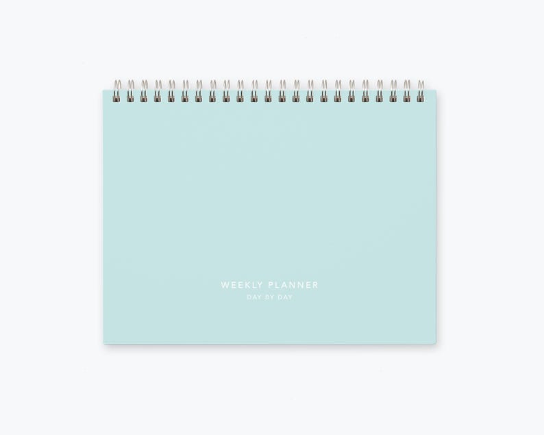 May include: A light blue weekly planner with a silver coil binding. The planner has the text "WEEKLY PLANNER DAY BY DAY" printed on the front.