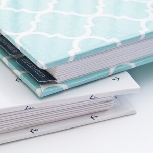 K&ouml;nnte beinhalten: Ein blaues und wei&szlig; gemustertes Hardcover-Journal mit einem wei&szlig;en R&uuml;cken und Laschen f&uuml;r jeden Monat. Die Laschen sind mit "November" und "Dezember" beschriftet. Das Journal liegt auf einem wei&szlig;en Journal mit einem blauen Anker-Design auf dem R&uuml;cken.