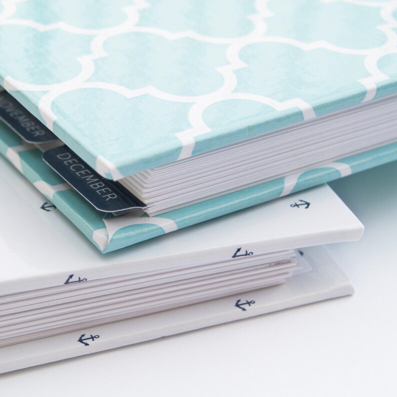 K&ouml;nnte beinhalten: Ein blaues und wei&szlig; gemustertes Hardcover-Journal mit einem wei&szlig;en R&uuml;cken und Laschen f&uuml;r jeden Monat. Die Laschen sind mit "November" und "Dezember" beschriftet. Das Journal liegt auf einem wei&szlig;en Journal mit einem blauen Anker-Design auf dem R&uuml;cken.