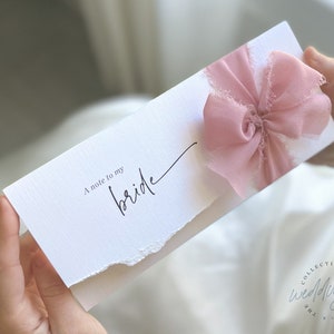 May include: A white card with a pink ribbon bow and the text "A note to my bride" written in black calligraphy.