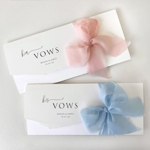 May include: Two white envelopes with the words "her vows" and "his vows" printed on them. The envelopes are tied with pink and blue ribbons.