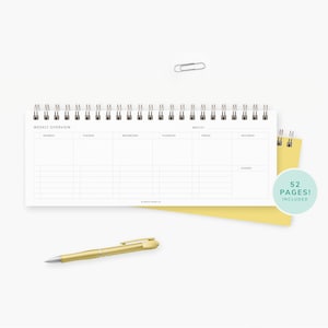 May include: A white spiral-bound weekly planner with a gold pen and a yellow notepad. The planner is open to a weekly overview page. A paper clip is above the planner. The planner includes 52 pages.
