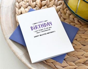 Op de afbeelding: Een witte verjaardagskaart met paarse tekst met de tekst "I didn't forget your BIRTHDAY. I just forgot to tell you that I remembered. HAPPY BELATED BIRTHDAY!" De kaart ligt op een blauwe envelop, rustend op een geweven dienblad.