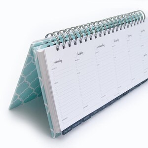 K&ouml;nnte beinhalten: Ein blauer Schreibtischkalender mit einem wei&szlig;en geometrischen Muster auf dem Umschlag. Der Kalender hat eine Spiralbindung und Laschen f&uuml;r jeden Monat des Jahres. Der Kalender ist auf der Seite f&uuml;r Januar, Februar, M&auml;rz und April ge&ouml;ffnet.