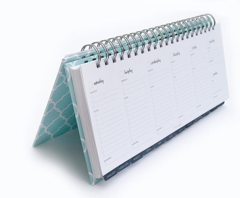 K&ouml;nnte beinhalten: Ein blauer Schreibtischkalender mit einem wei&szlig;en geometrischen Muster auf dem Umschlag. Der Kalender hat eine Spiralbindung und Laschen f&uuml;r jeden Monat des Jahres. Der Kalender ist auf der Seite f&uuml;r Januar, Februar, M&auml;rz und April ge&ouml;ffnet.