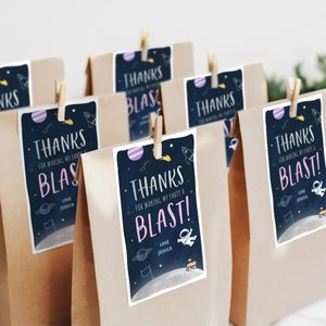 Pode incluir: Um conjunto de sacos de papel castanho com etiquetas de agradecimento anexas. As etiquetas apresentam um design com tema espacial com planetas, estrelas e um foguete. O texto nas etiquetas diz "Obrigado por fazer da minha festa uma explos&atilde;o!" com um cora&ccedil;&atilde;o rosa e o nome Jessica.