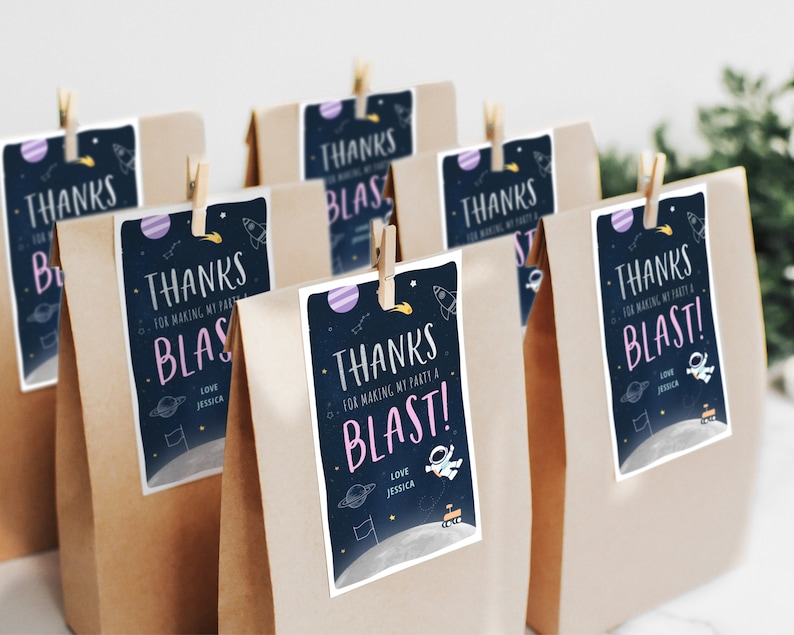 Pode incluir: Um conjunto de sacos de papel castanho com etiquetas de agradecimento anexas. As etiquetas apresentam um design com tema espacial com planetas, estrelas e um foguete. O texto nas etiquetas diz "Obrigado por fazer da minha festa uma explos&atilde;o!" com um cora&ccedil;&atilde;o rosa e o nome Jessica.