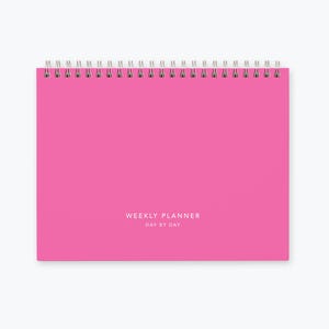 May include: A pink weekly planner with a spiral binding and the text "WEEKLY PLANNER DAY BY DAY" printed on the front cover.