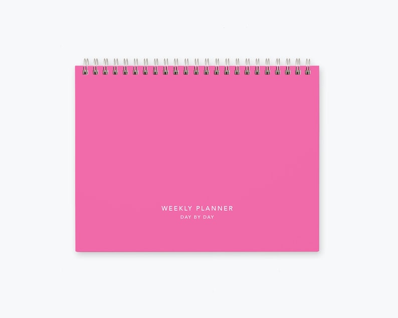 May include: A pink weekly planner with a spiral binding and the text "WEEKLY PLANNER DAY BY DAY" printed on the front cover.