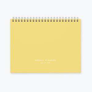May include: A yellow weekly planner with a spiral binding and the text "WEEKLY PLANNER DAY BY DAY" printed on the front.