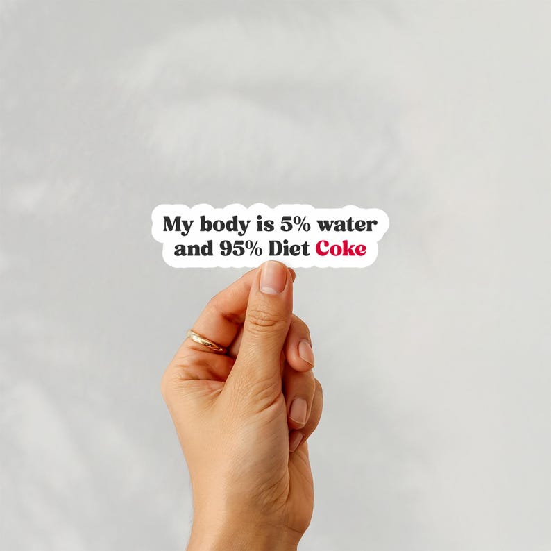 May include: A white sticker with the text "My body is 5% water and 95% Diet Coke" in black and red font. The sticker is held by a hand with a gold ring.