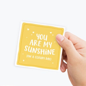 May include: A yellow card with the text "You are my sunshine (on a cloudy day)" in white lettering. The card has a white border and small white hearts and stars scattered around the text.
