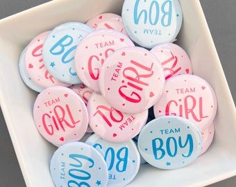 Könnte beinhalten: Eine Sammlung runder Buttons in einer weißen Schale. Die Buttons sind rosa und hellblau, mit den Wörtern "Team Girl" und "Team Boy" in Schriftform. Einige Buttons haben Sterne und Herz-Akzente.