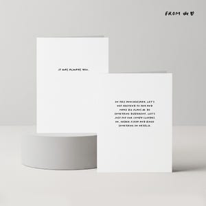 May include: Two white greeting cards with black text. One card reads "IT WAS ALWAYS YOU." The other card says, "ON THIS ANNIVERSARY, LET'S NOT PRETEND TO TRY AND MAKE BIG PLANS OR DO SOMETHING DIFFERENT..." The cards are on a white pedestal.
