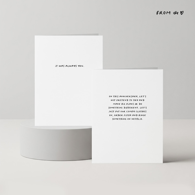May include: Two white greeting cards with black text. One card reads "IT WAS ALWAYS YOU." The other card says, "ON THIS ANNIVERSARY, LET'S NOT PRETEND TO TRY AND MAKE BIG PLANS OR DO SOMETHING DIFFERENT..." The cards are on a white pedestal.