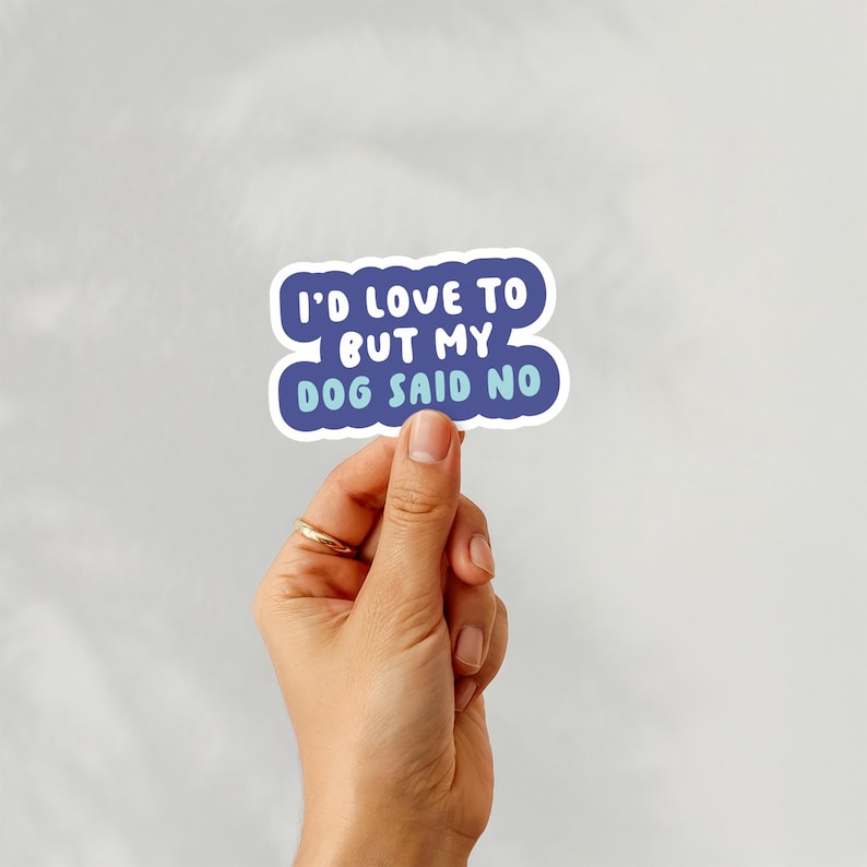 May include: A white sticker with blue text that reads "I'D LOVE TO BUT MY DOG SAID NO." The text is in a playful font and is outlined in white. The sticker is held by a hand with a gold ring.