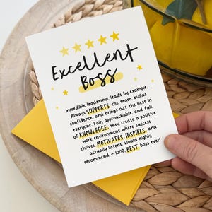 May include: A white card with the words "Excellent Boss" in black script, accented with yellow stars. The card is held over a yellow envelope and a woven tray. Text on the card praises leadership qualities.