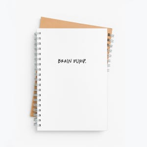 May include: A white spiral bound notebook with the words "BRAIN DUMP." written in black marker on the front cover. The notebook is sitting on a brown paper background.
