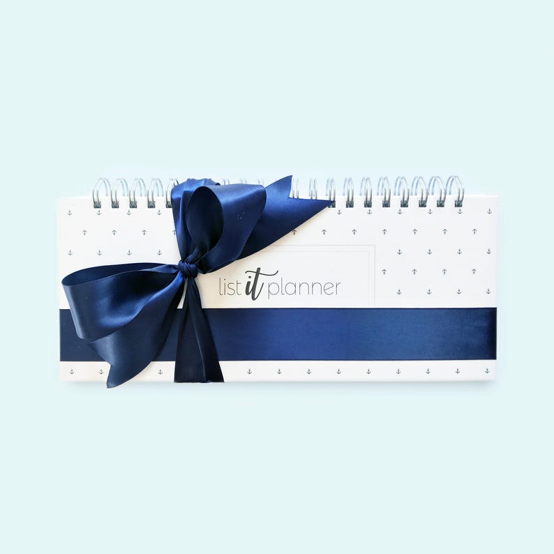May include: A white planner with a navy blue ribbon tied in a bow. The planner has a white background with a repeating pattern of small anchors. The planner has the text "list it planner" printed on the front.