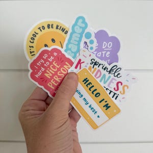 Puede incluir: Una colecci&oacute;n de pegatinas coloridas con mensajes y frases positivas. Las pegatinas incluyen frases como "It's cool to be kind", "Sprinkle Kindness" y "Hello I'm trying my best". Las pegatinas se sostienen en una mano.