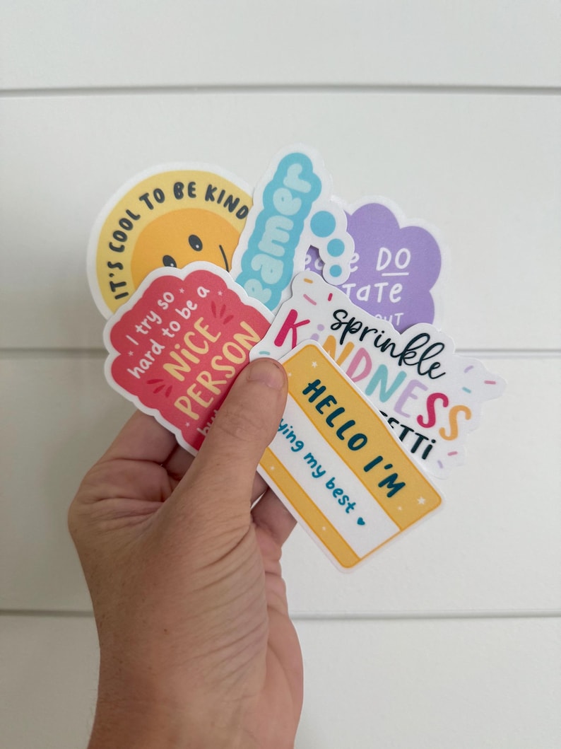Puede incluir: Una colecci&oacute;n de pegatinas coloridas con mensajes y frases positivas. Las pegatinas incluyen frases como "It's cool to be kind", "Sprinkle Kindness" y "Hello I'm trying my best". Las pegatinas se sostienen en una mano.